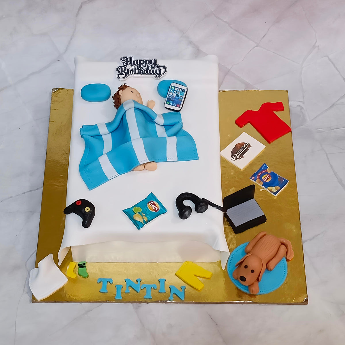 A custom fondant cake shaped like a child's bed, decorated with a blue and white striped blanket, a sleeping fondant figure, and various themed accessories including a phone, game controller, snacks, headphones, a laptop, a dog, and 'TINTIN' written at the bottom. 