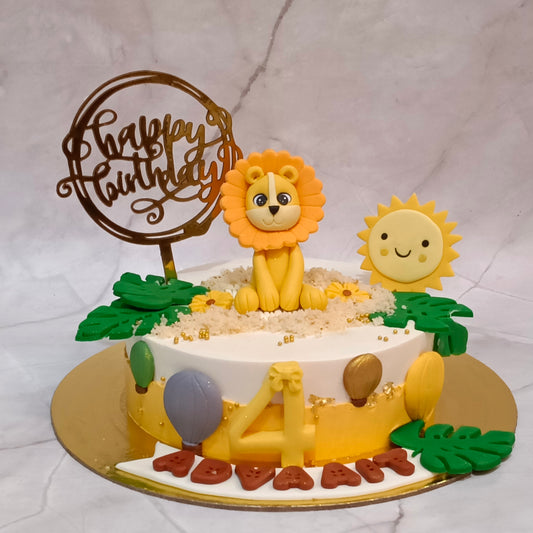 A round birthday cake decorated with a yellow lion figurine, a smiling sun, green leaves, and a gold 'Happy Birthday' topper, presented on a gold cake board against a light marble-patterned backdrop.