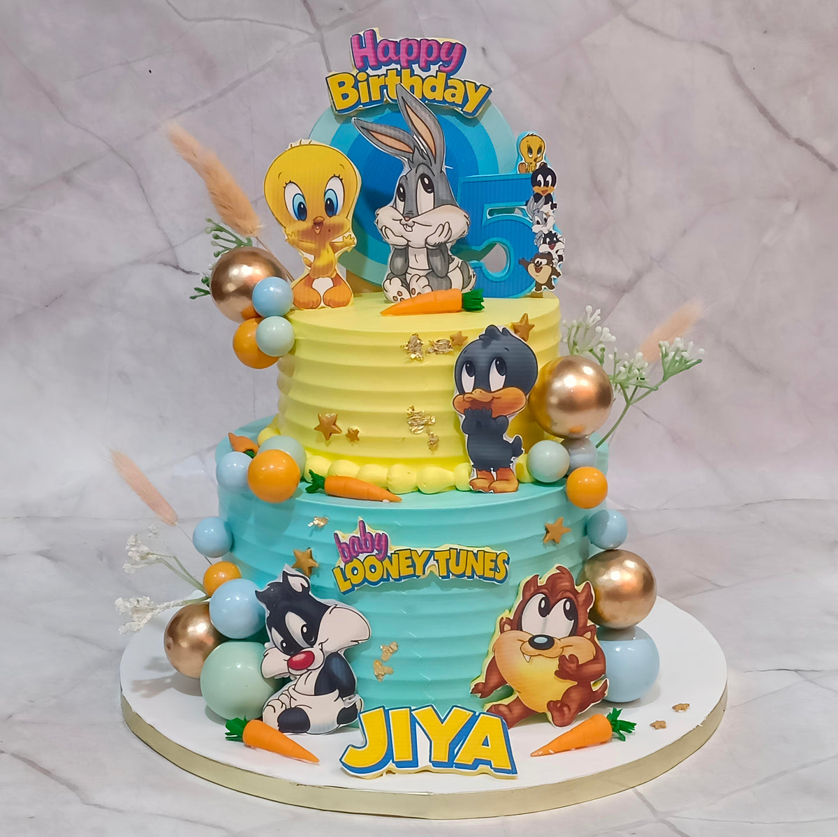 A two-tier Looney Tunes birthday cake decorated with cartoon characters including Tweety, Bugs Bunny, Daffy Duck, Tweety, Sylvester, Tweety, and the name 'JIYA' on the front