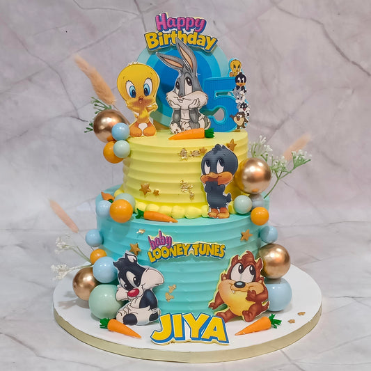 A two-tier Looney Tunes birthday cake decorated with cartoon characters including Tweety, Bugs Bunny, Daffy Duck, Tweety, Sylvester, Tweety, and the name 'JIYA' on the front