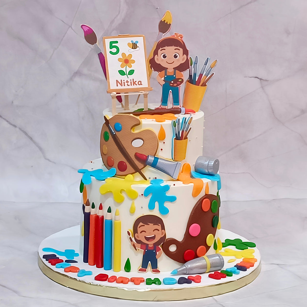 A decorated two-tier birthday cake for an artist, featuring fondant paint splatters, a large fondant paint palette, colored pencils, paint tubes, and cartoon character toppers of children painting