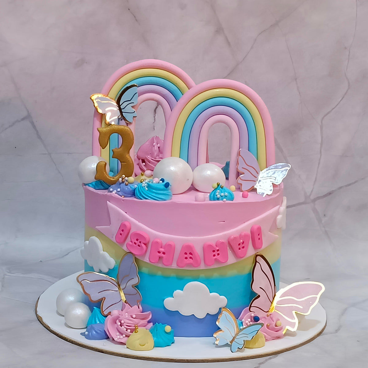 Rainbow Butterfly 3rd Birthday Cake for a girl, decorated with two rainbows, clouds, fondant balls, faux butterflies and an age 3 tag.