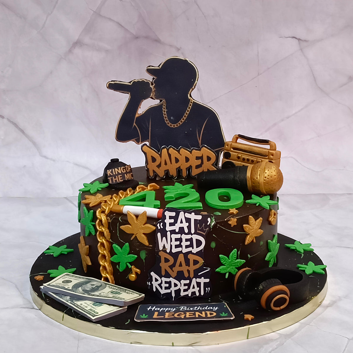 A custom chocolate cake decorated with a black silhouette of a rapper holding a microphone, gold-colored toppers, green dollar sign dollar bills, cannabis leaves, and the text '420', 'EAT WEED REAP RAP', and 'Happy Birthday Legend'.