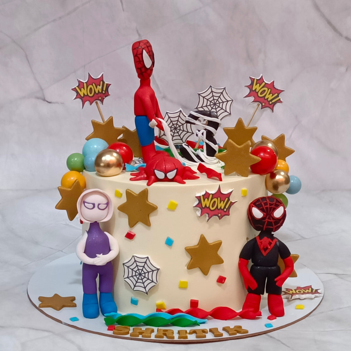 A custom Spider-Man themed birthday cake decorated with Spider-Man and Spider-Gwen figurines, gold stars, spiderweb motifs, and 'WOW!' toppers 