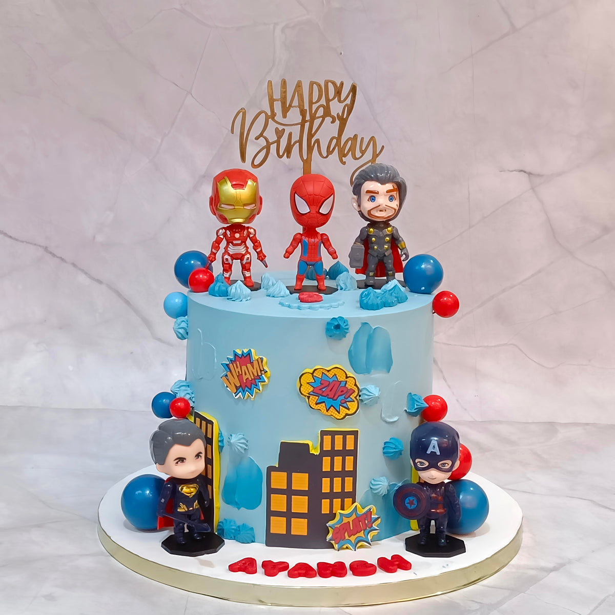A custom Marvel-themed birthday cake with a light blue frosting base, decorated with character toppers including Iron Man, Spider-Man, Captain America, and Thor, along with comic book speech bubbles and city silhouettes. The cake is topped with a gold 'Happy Birthday' topper and stands on a round white base.