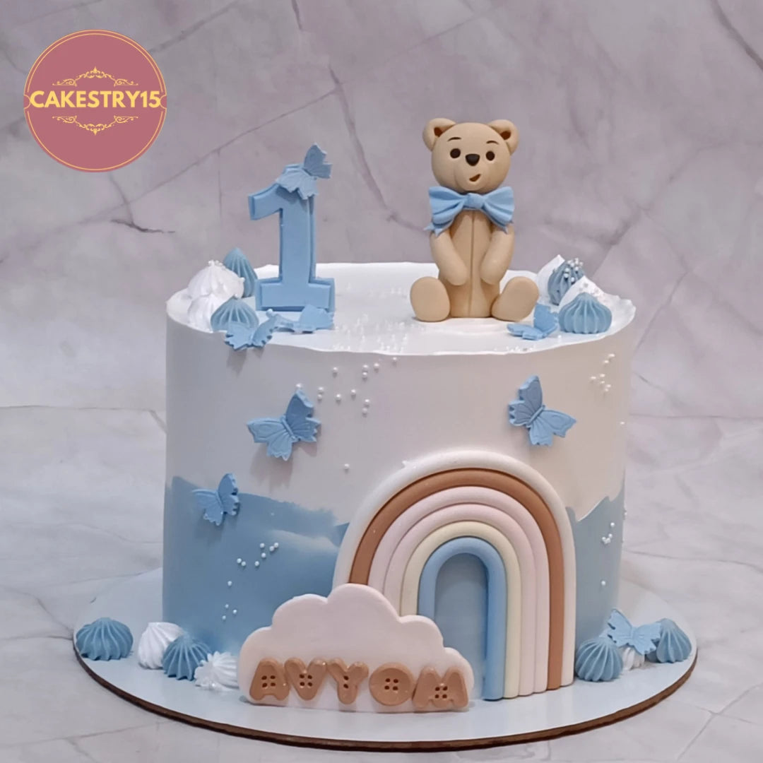 Eggless teddy and rainbow kids birthday cake with clouds and butterflies, a premium theme cake Noida design for first birthday celebrations.