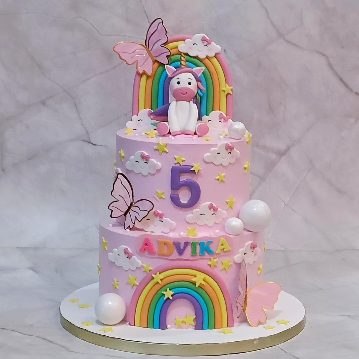 Two-tier unicorn birthday cake with rainbows, butterflies, clouds, faux balls and stars, personalised with ADVIKA and number 5.