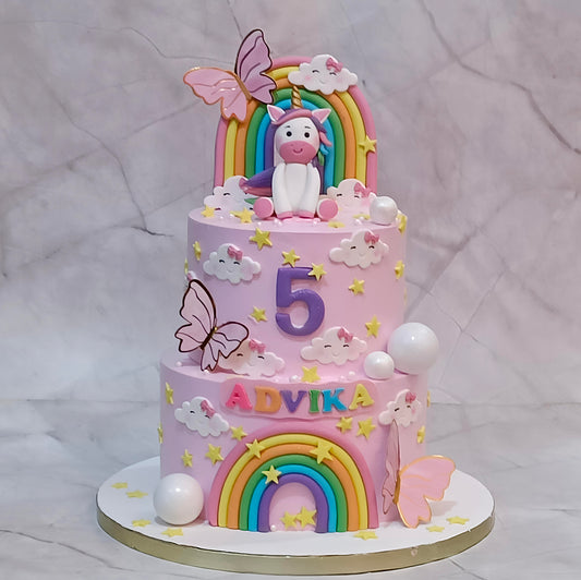 Two-tier unicorn birthday cake with rainbows, butterflies, clouds, faux balls and stars, personalised with ADVIKA and number 5.
