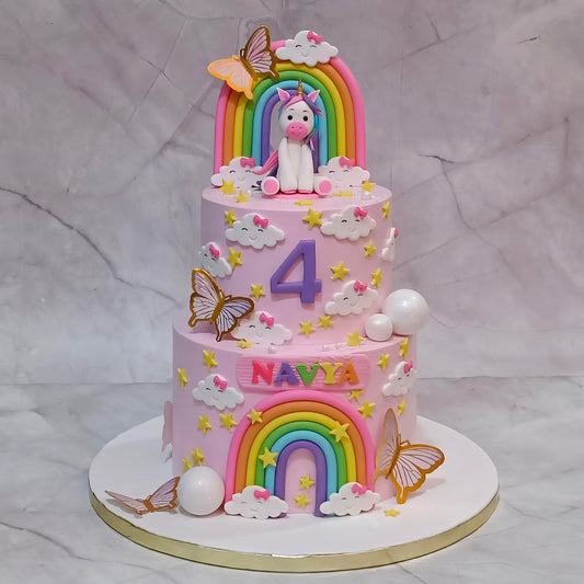 A pink two-tier birthday cake decorated with fondant unicorns, rainbows, clouds, butterflies, stars, and the name 'NAVYA' on the front, displayed on a white round cake board against a light marble-patterned backdrop.