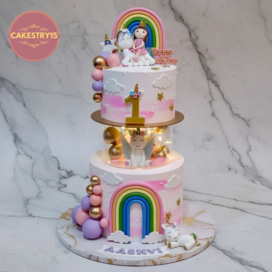 A three-tier pastel unicorn birthday cake with rainbow decorations, gold balloons, and unicorn toppers, displayed on a marble surface with a marble background. A 'CAKESTRY15' logo is visible in the top left corner.