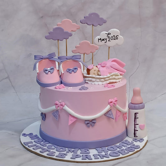 A pink and lavender baby shower cake decorated with fondant baby shoes, a sleeping baby figure, pink and purple clouds, a baby bottle, and a sign reading '2nd May 2026'. The cake sits on a white base with the text 'WELCOME HOME LITTLE PRINCESS' and is photographed against a light gray marble-patterned backdrop.