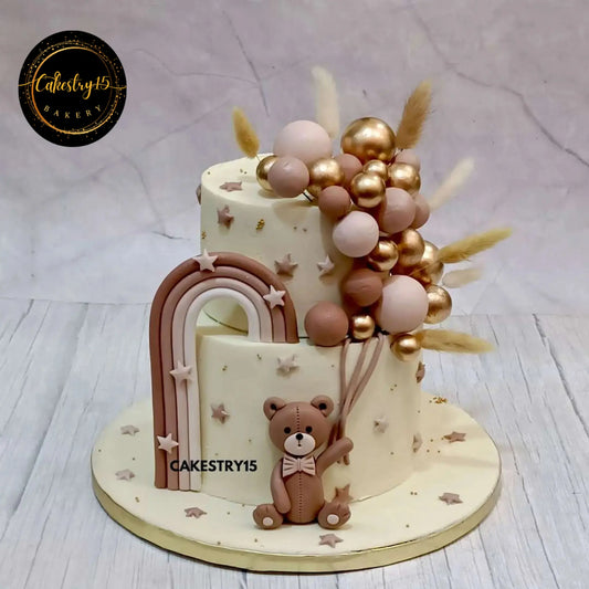 A 2-tier chocolate welcome baby cake featuring a teddy at the base holding faux balloon clusters, finished with milk chocolate ganache.