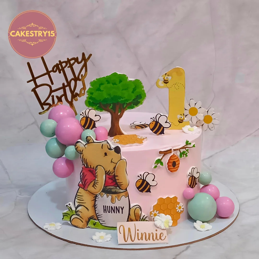 A pink round birthday cake decorated with Winnie the Pooh elements, including a bear figure, honey pot, bees, flowers, a green tree, a yellow number 1 topper, and a 'Happy Birthday' topper. The cake is displayed on a marble-patterned background with a 'CAKESTRY15' logo in the top left corner.